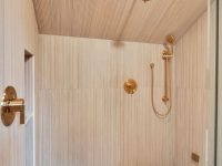 Modern walk-in shower with light wood paneled walls, marble floor, brass fixtures, and glass enclosure
