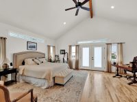 Spacious master bedroom with wood flooring, large bed, seating area, glass doors, ceiling fan, and natural light from multiple windows