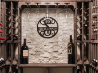 Wine cellar display with brick wall, decorative circular metal sign, wine bottles on wooden shelving, and ambient lighting